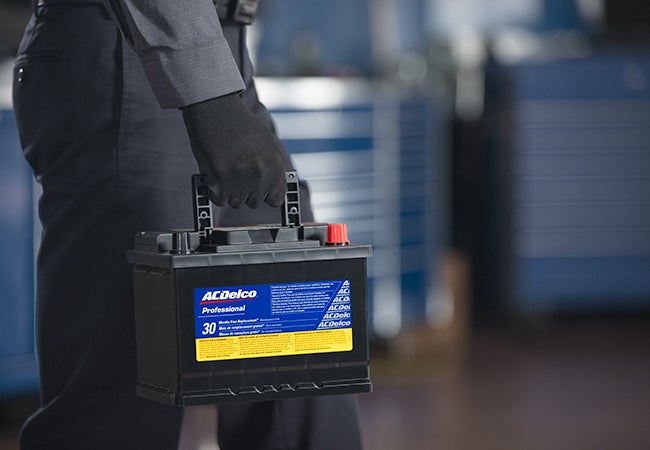 Close-up of a person in black gloves and a navy uniform carrying an AC Delco Professional car battery.