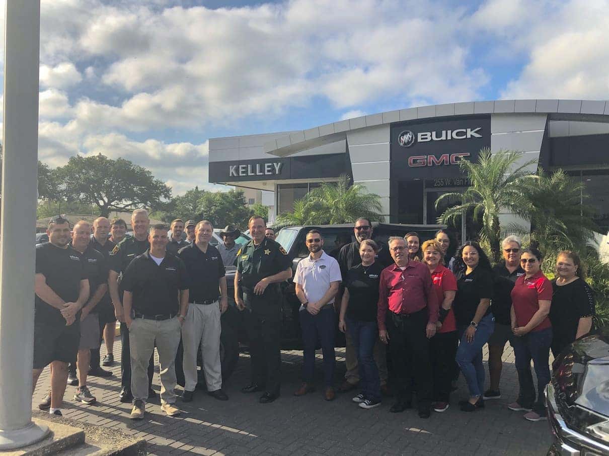 A large group of men and women, including law enforcement and civilians, pose for a photo in front of a Kelley car dealership.