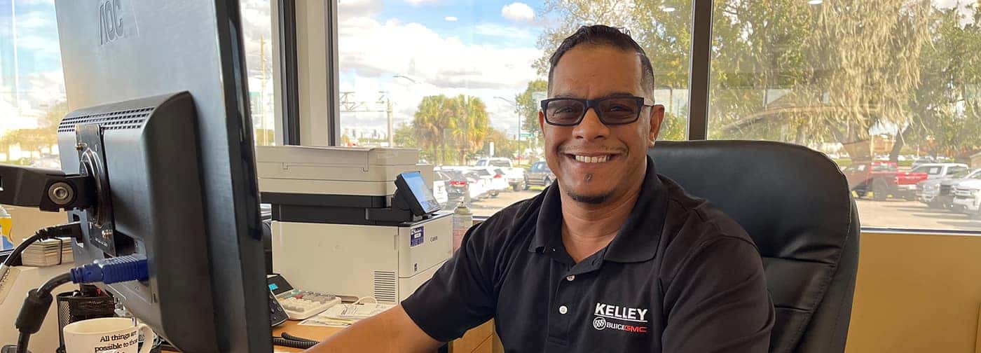 Man in a black polo shirt with KELLEY BUICK GMC logo, smiling at a desk in an office with large windows.
