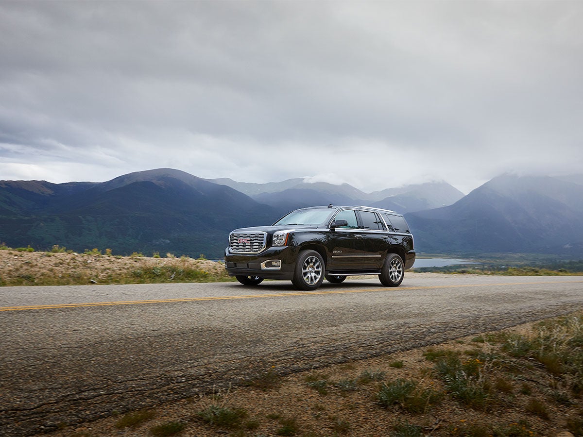 Dark gray SUV on a road with mountains, a lake, and cloudy skies in the background.
