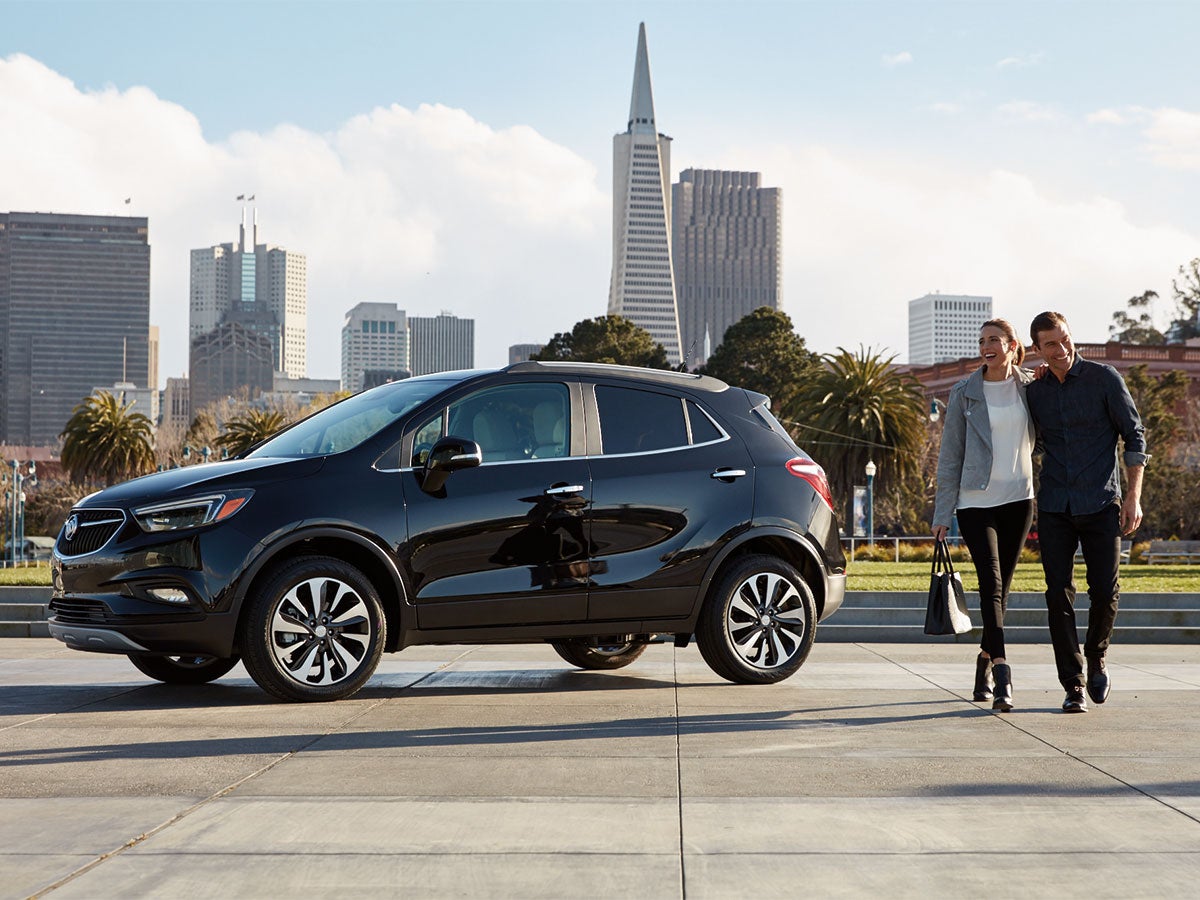 A black Buick Encore with a smiling couple walking beside it in front of a city skyline with palm trees.