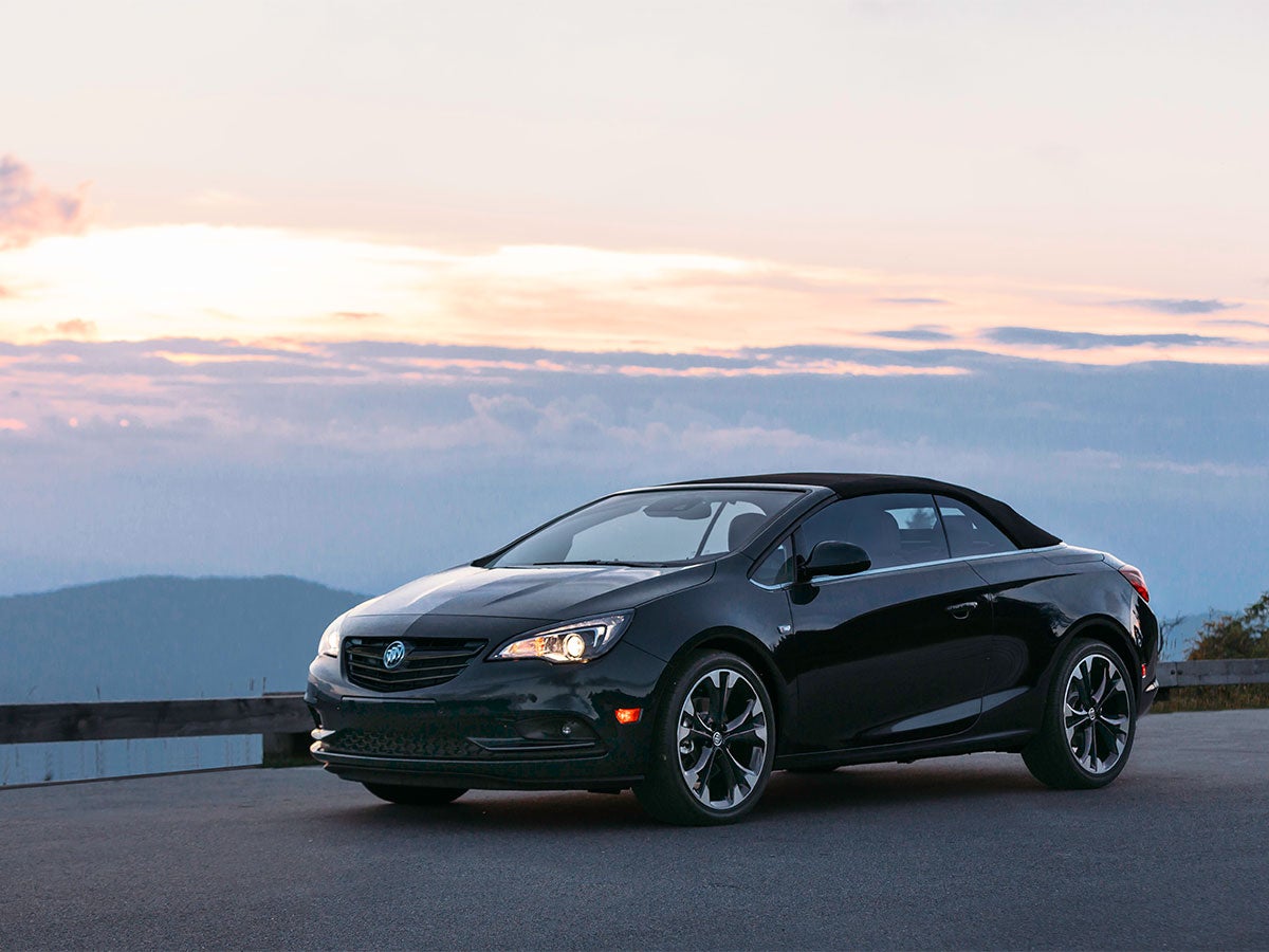 Black convertible car with its top up, parked with a sunset sky over distant mountains in the background.