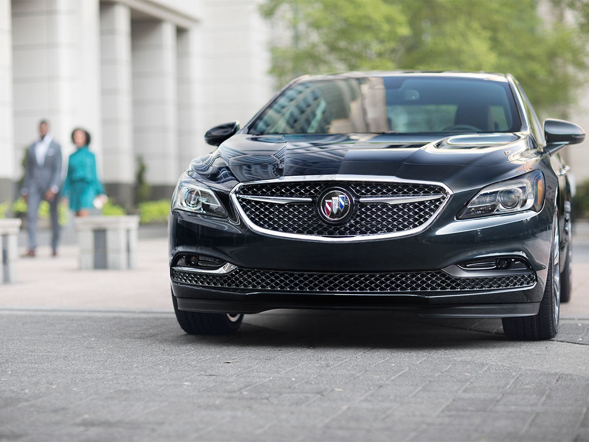 Front view of a dark gray Buick car parked on asphalt, with two people walking in the background.