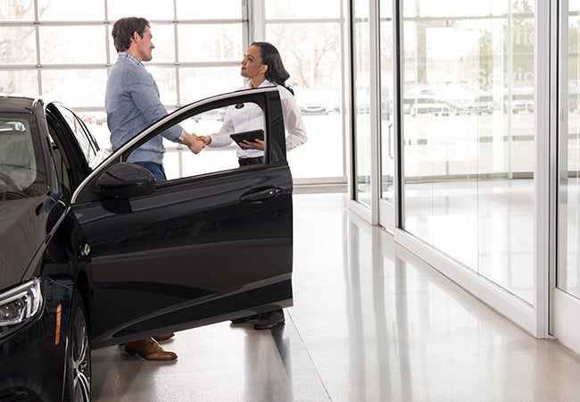 A man and woman shake hands in a car dealership next to a black car with its door open.