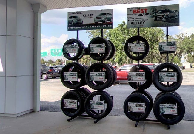 Outdoor tire display with varying prices shown, under Kelley banners featuring cars.