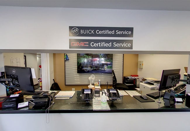 A service reception counter with two monitors, phones, and a printer on a black countertop; above are BUICK Certified Service and GMC Certified Service signs.