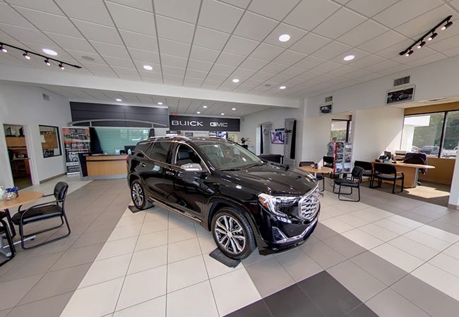 A black GMC SUV displayed in a car dealership showroom with a reception desk and customer seating areas.