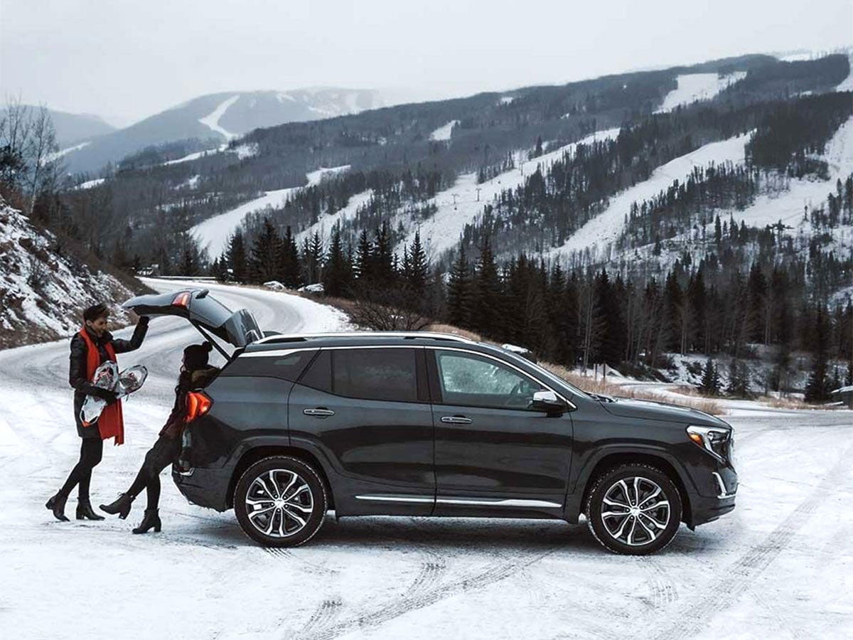 Two people unload bags from a dark grey SUV on a snowy mountain road, with ski slopes in the background.