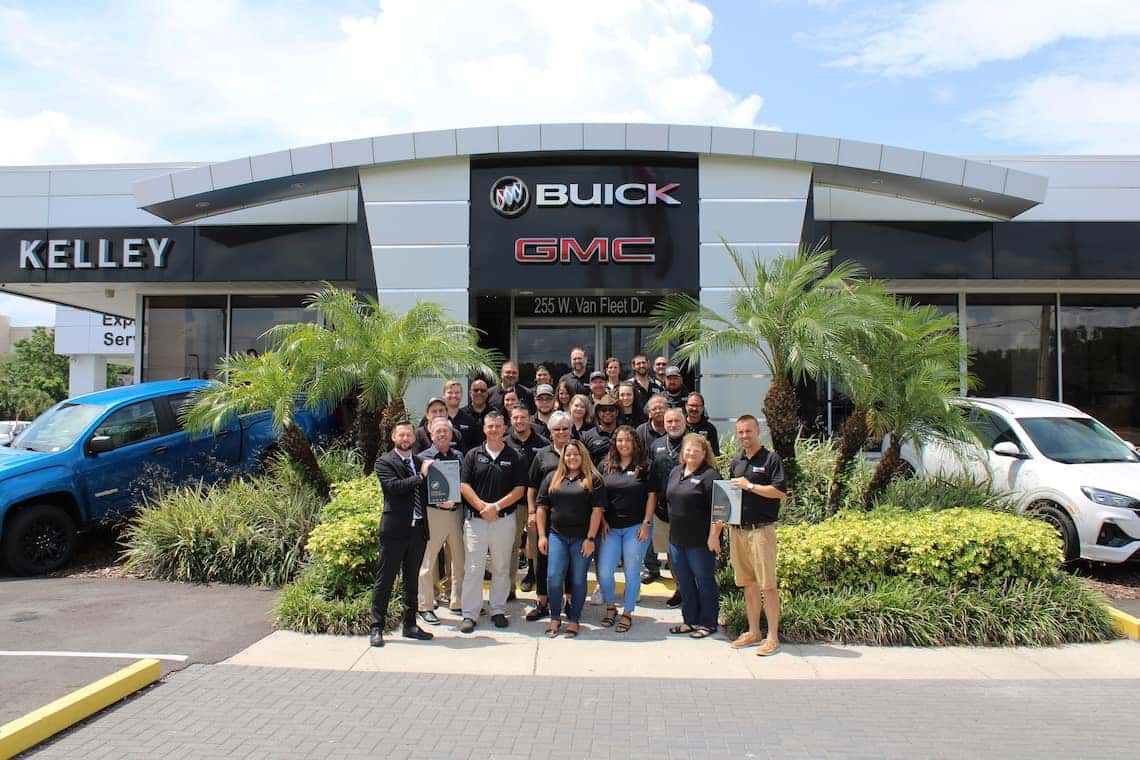 Group of dealership employees smiling and holding awards in front of a Buick GMC dealership building with palm trees.