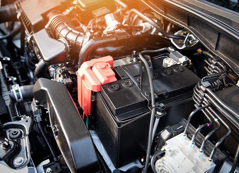 Close-up of a car engine bay, showing the black battery with a red terminal cover and surrounding mechanical parts.